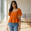 A woman smiling outdoors in a garden, wearing a bright orange puff-sleeve peplum top and light blue jeans.