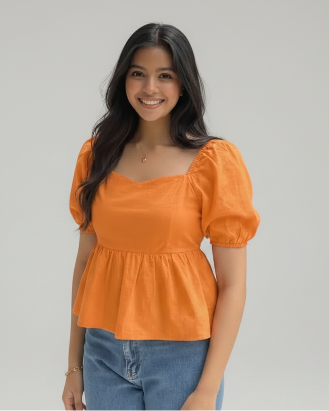A woman wearing a bright orange puff-sleeve peplum top and blue jeans stands outdoors in a garden setting with green foliage and yellow flowers.