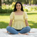 A woman sits cross-legged on a white blanket in a sunny park, wearing a pale yellow puff-sleeve top with a square neckline and blue jeans.