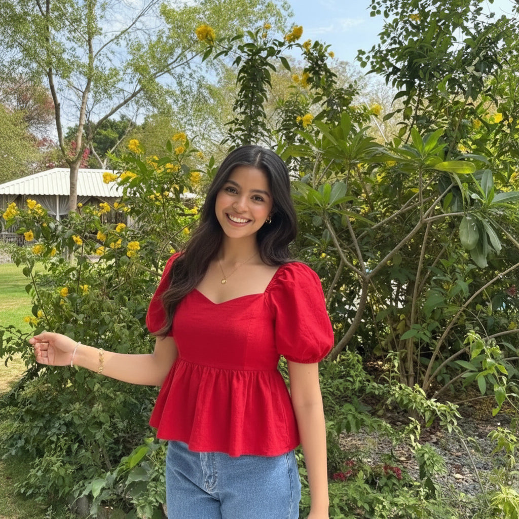 A woman smiling outdoors in a bright red puff-sleeve top and light blue jeans, standing in front of lush green garden foliage with yellow flowers.