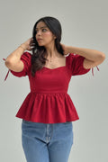 A woman wearing a red puff-sleeve peplum top and light blue jeans, standing outdoors in a garden with green foliage and yellow flowers.