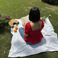 A woman viewed from behind sits on a white blanket on a grassy lawn, wearing a bright red puff-sleeve top with a smocked back and peplum hem. She is holding an open book, with a woven hat, two oranges, and a small bouquet of yellow and white flowers nearby.
