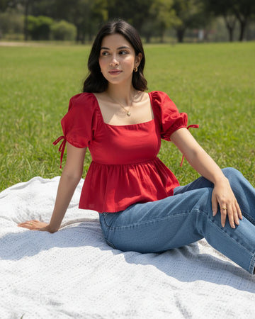 A woman wearing a bright red puff-sleeve top with a square neckline and side tie details, paired with blue jeans, sitting outdoors on a white blanket on grass.