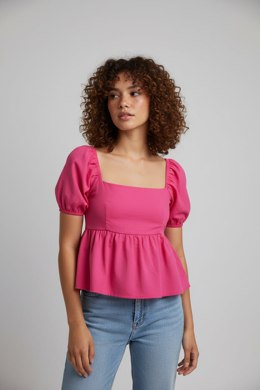 A woman wearing a bright pink peplum top and blue jeans stands outdoors in front of lush green foliage and yellow flowers.