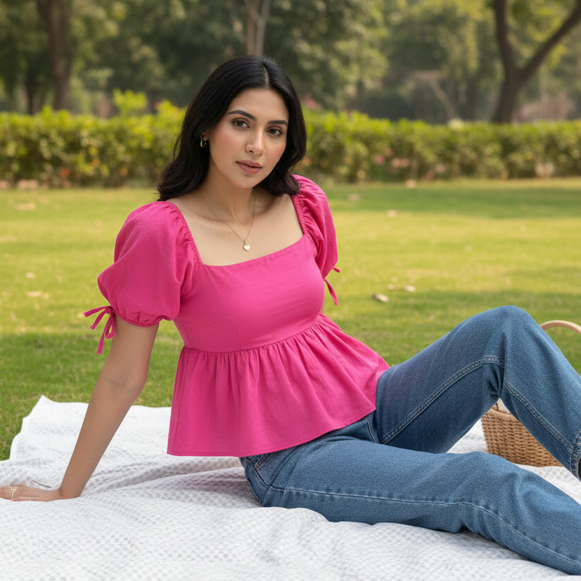 A woman wearing a bright pink puff-sleeve top with a square neckline and gathered peplum hem, paired with blue jeans, sitting outdoors on grass.