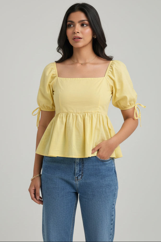 A woman modeling a pale yellow puff-sleeve blouse with a square neckline and tie details at the sleeves, paired with blue jeans, against a solid light gray background.