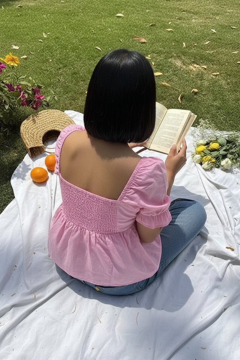 A woman viewed from behind wearing a pink puff-sleeve smocked blouse and blue jeans, sitting on a white blanket on grass with an open book, flowers, oranges, and a straw hat nearby.