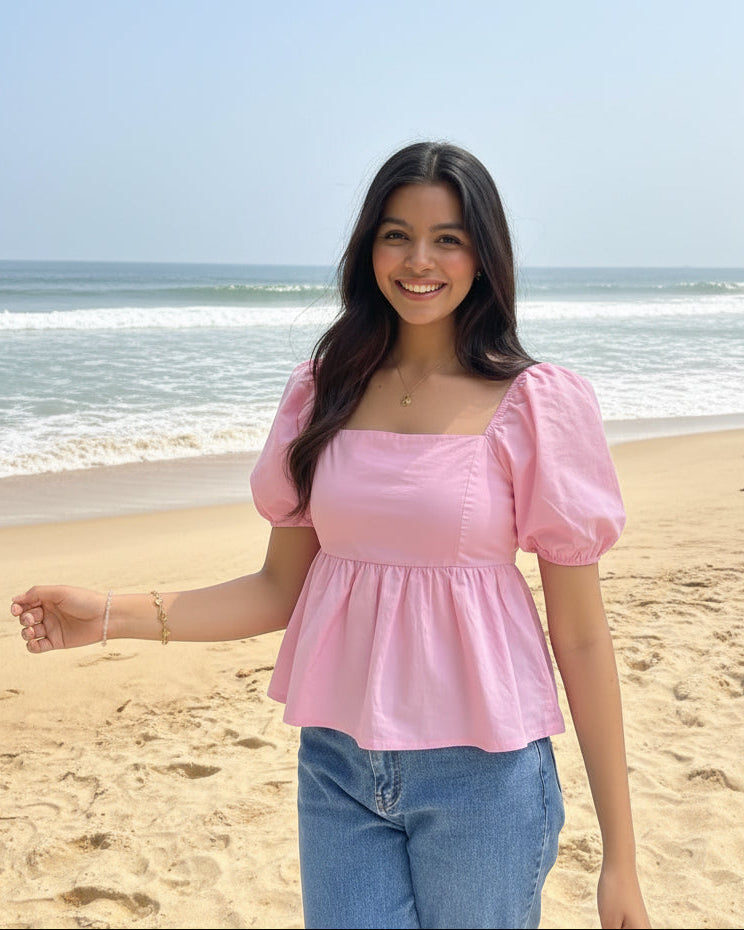 A woman smiling outdoors in a garden, wearing a light pink puff-sleeve peplum top and blue jeans.