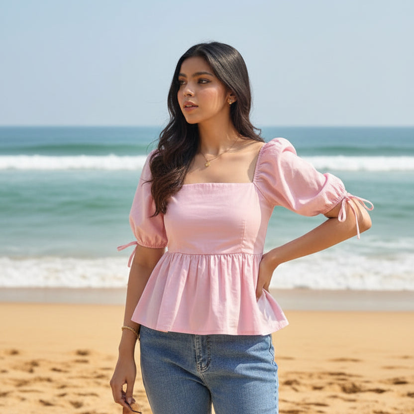 A woman wearing a light pink peplum top with puff sleeves and a square neckline, paired with blue jeans, standing on goa beach