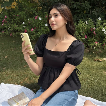 A woman wearing a black puff-sleeve top with a square neckline and peplum hem, paired with blue jeans, sitting outdoors on a white blanket on grass.