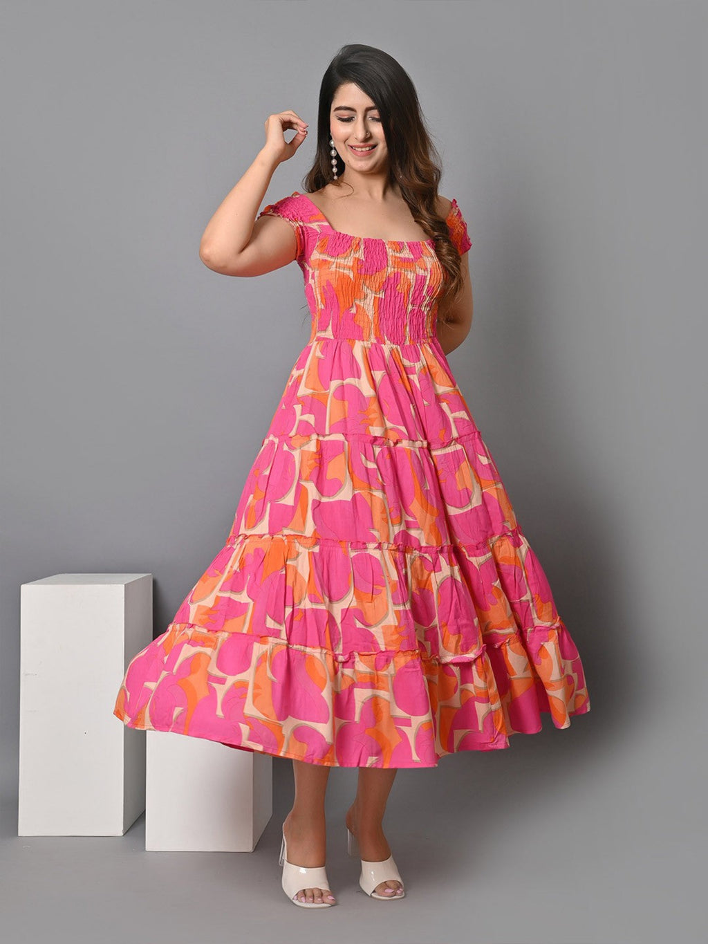 A woman wearing a pink and orange geometric print A-line dress with short sleeves and a flared hem, standing next to white geometric blocks.
